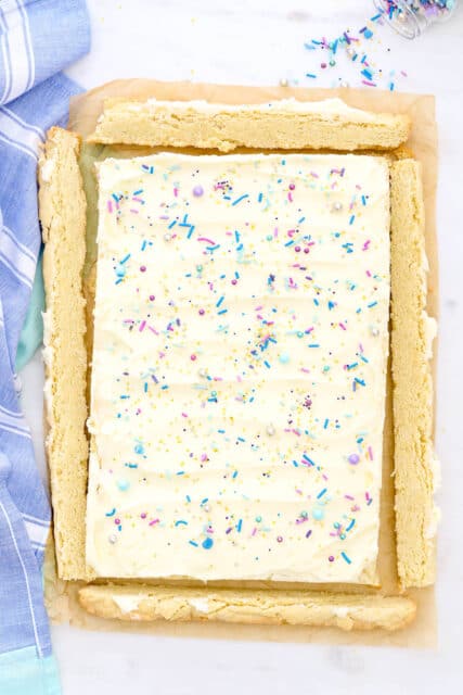 Frosted sugar cookie bars topped with rainbow sprinkles on a piece of parchment paper.