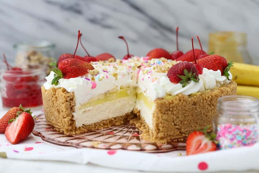 A whole no-bake banana split pie on a pink pie plate with a slice missing showing the inside of the pie.