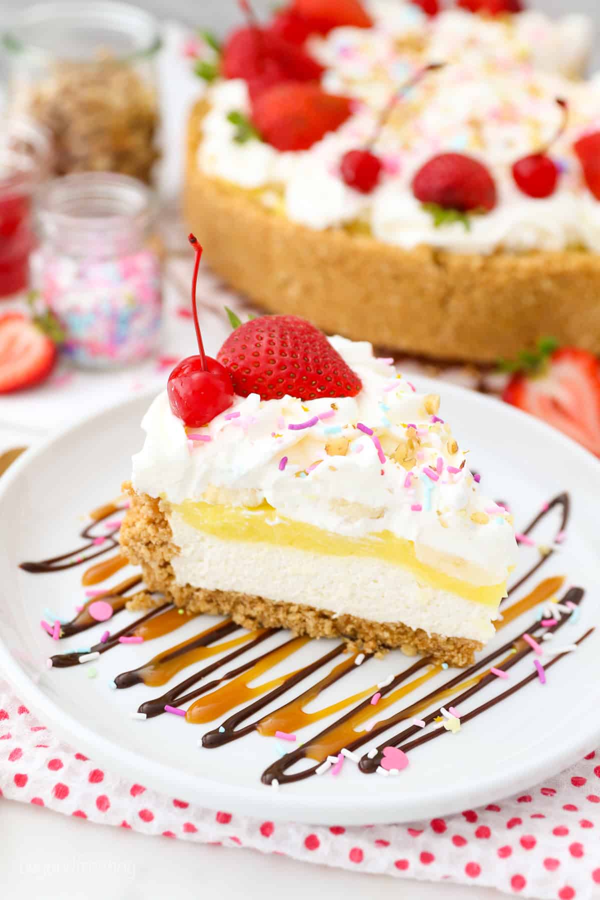 Banana split pie has all the flavors you love in a banana split, with creamy no-bake cheesecake, banana pudding, and all the sundae toppings! A slice of banana split pie topped with whipped cream, maraschino cherries, and sprinkles on a plate with the rest of the no-bake pie in the background.