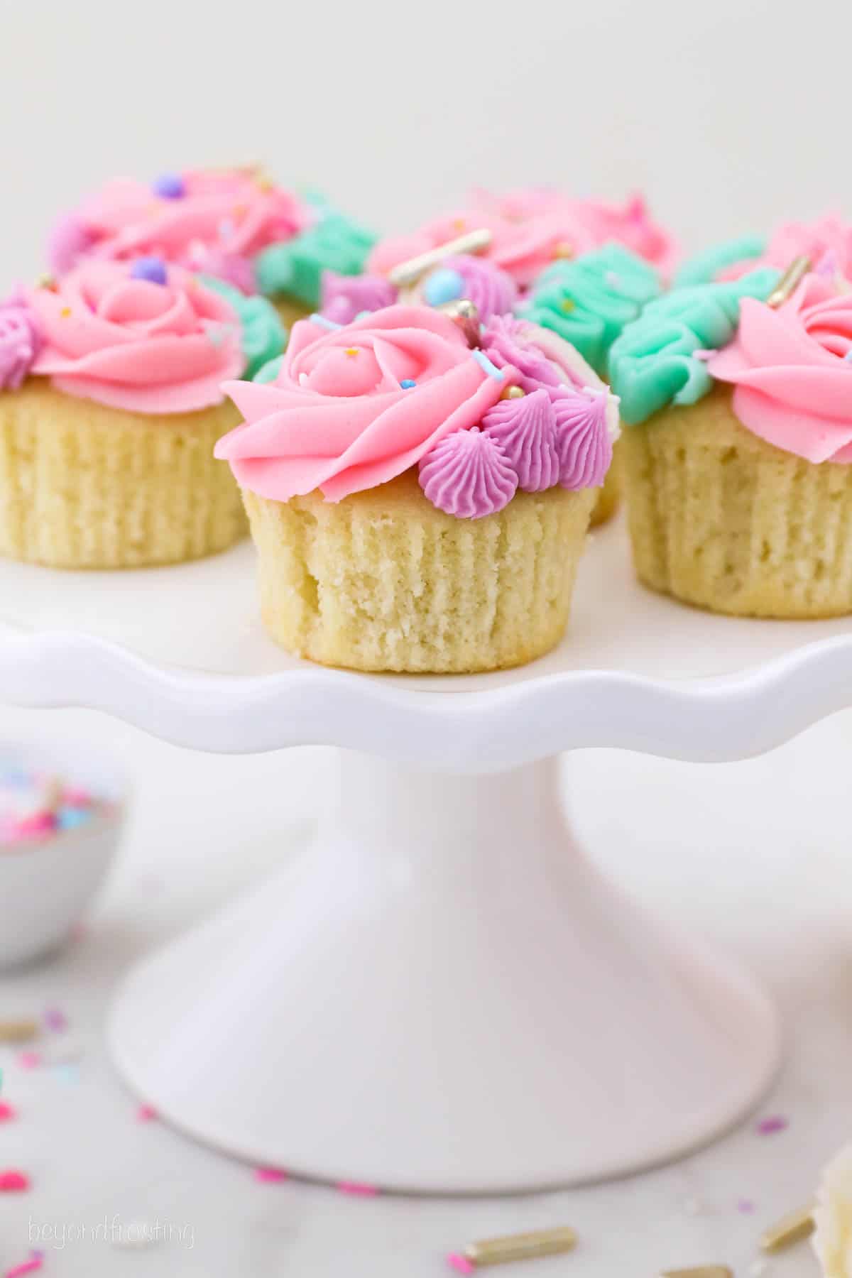 Brightly frosted unicorn cupcakes topped with sprinkles on a white cake stand.