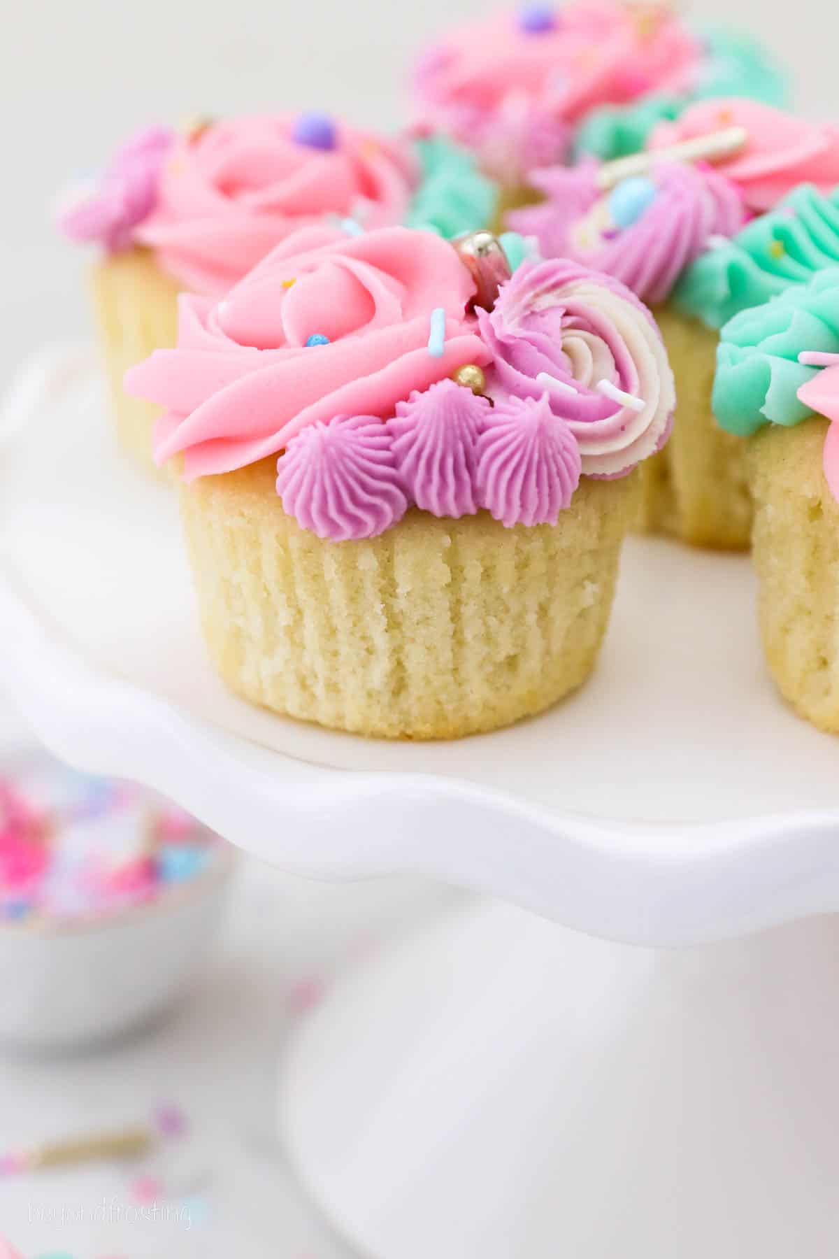 Close up of frosted unicorn cupcakes topped with sprinkles on a white cake stand.