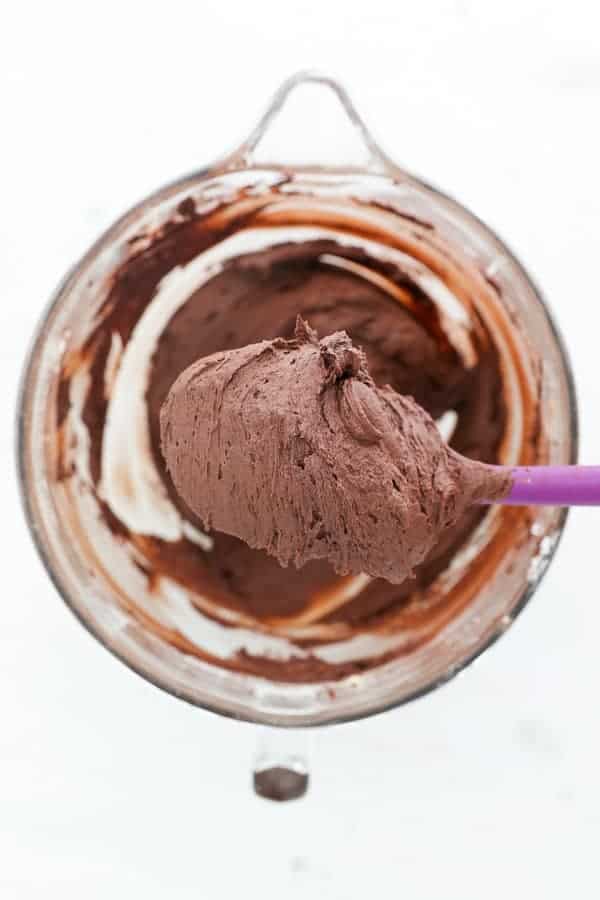 A spatula filled chocolate chocolate frosting sitting overtop of a glass mixing bowl