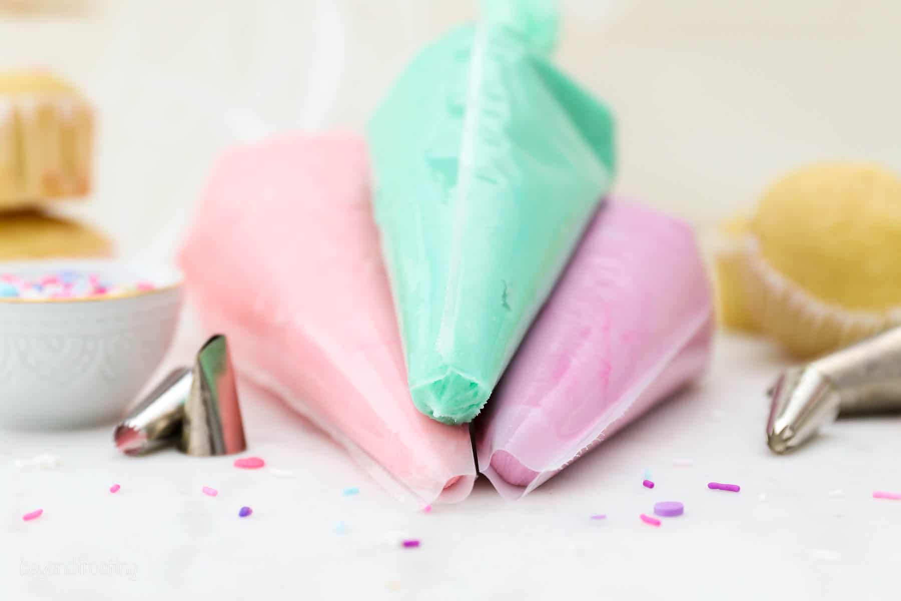 Three piping bags filled with pink, teal, and purple buttercream frosting stacked on a countertop next to piping tips.
