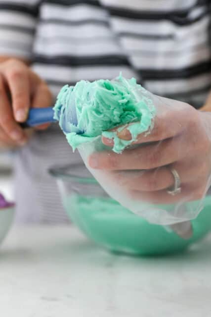 One hand holding a piping bag while the other uses a spatula to fill it with teal frosting.