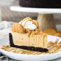 A close up shot of a slice of fluffy peanut butter pie on a white rimmed plate. There's a cake stand in the background with the pie sitting on it. It has an Oreo crust.