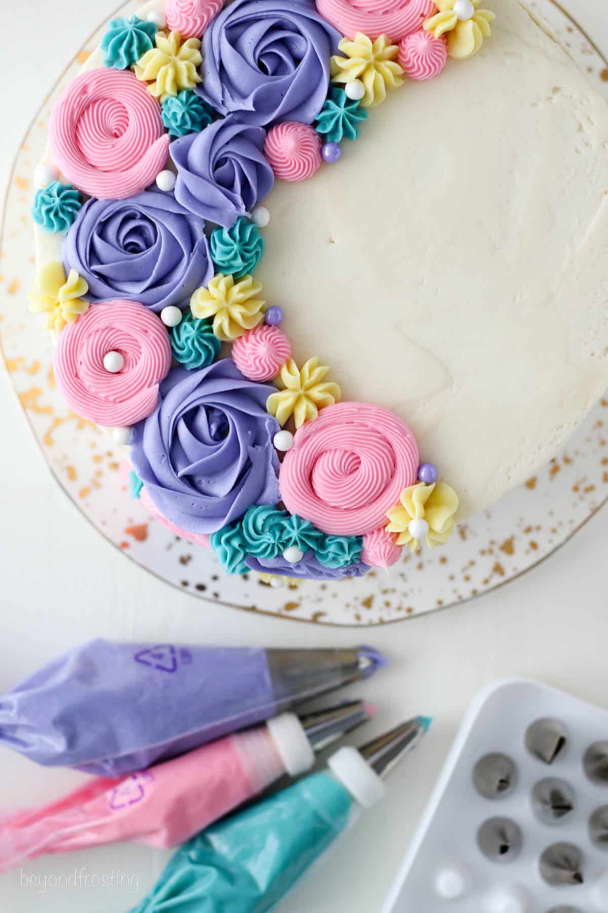 Overhead view of three piping bags with purple, pink, and light turquoise buttercream frosting next to a cake decorated with piped frosting flowers.