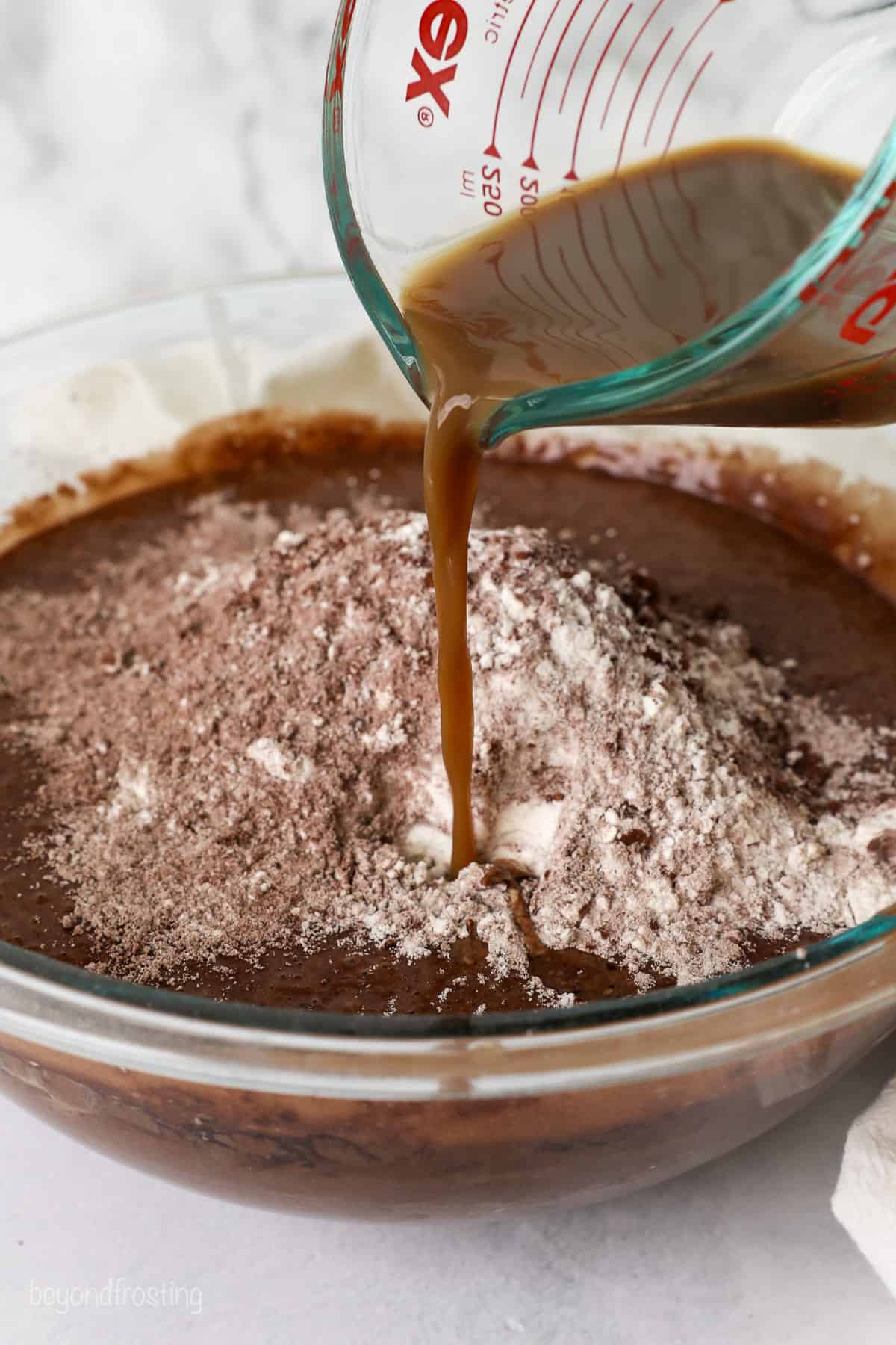 Brewed coffee is poured into the combined cake batter ingredients in a glass bowl.