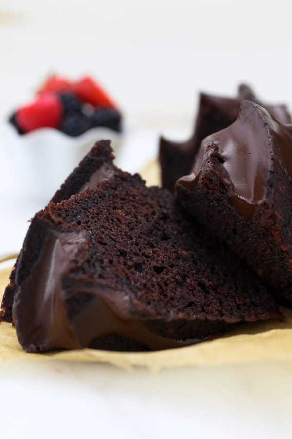 A couple slices of chocolate bundt cake laying on a piece of brown parchment paper and a bowl of fruit blurred out in the background