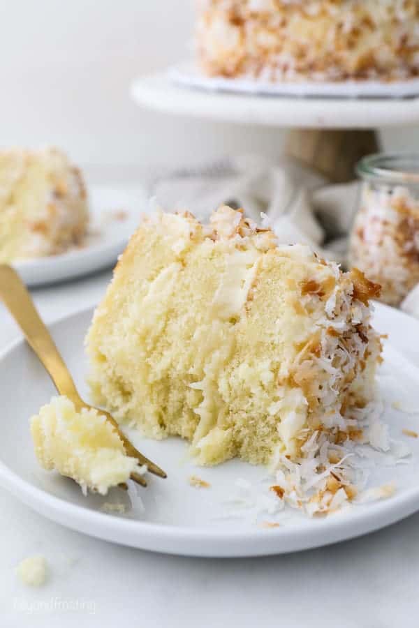 A slice of coconut cake laying on a white plate with a few bites missing, a gold fork laying on the plate has a bite of cake on it