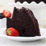 A slice of chocolate bundt cake on a white plate next to a strawberry.