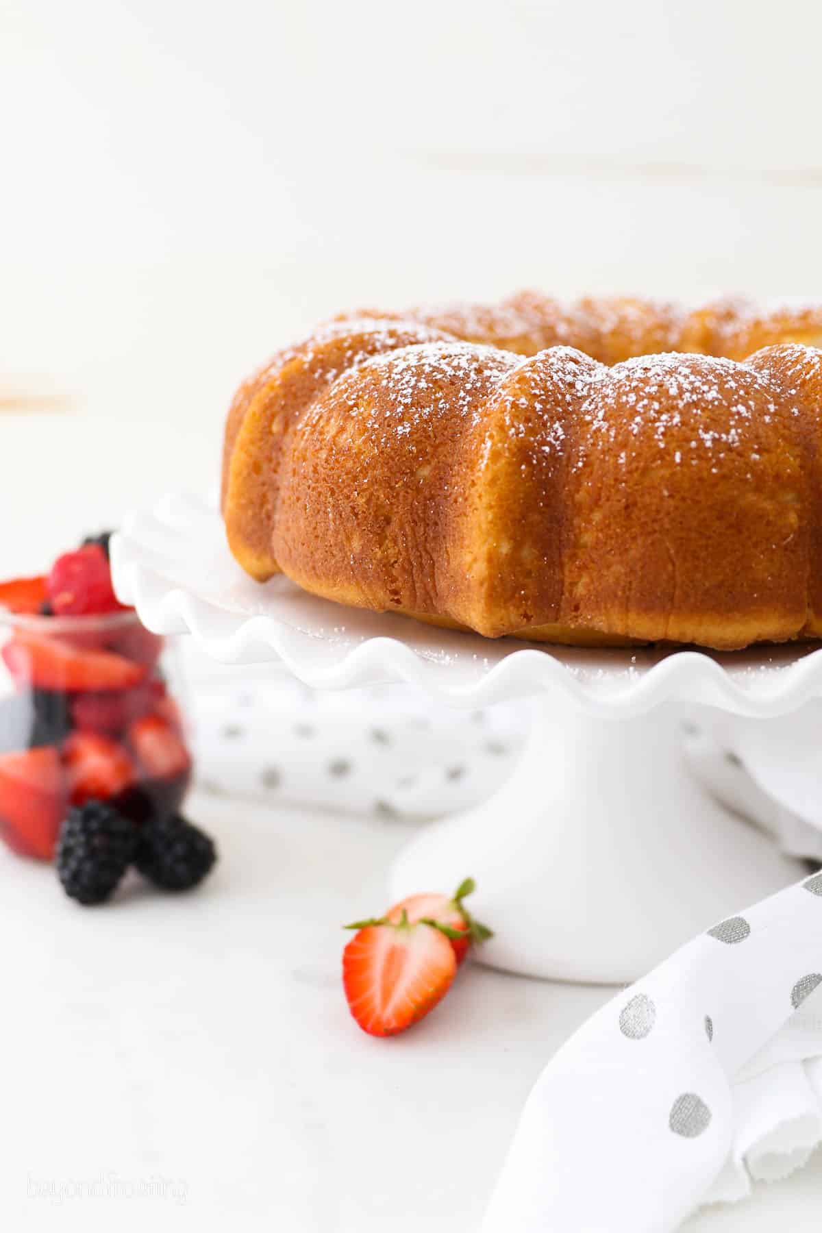 Vanilla bundt cake on a white cake stand on a countertop next to a jar of fresh berries.