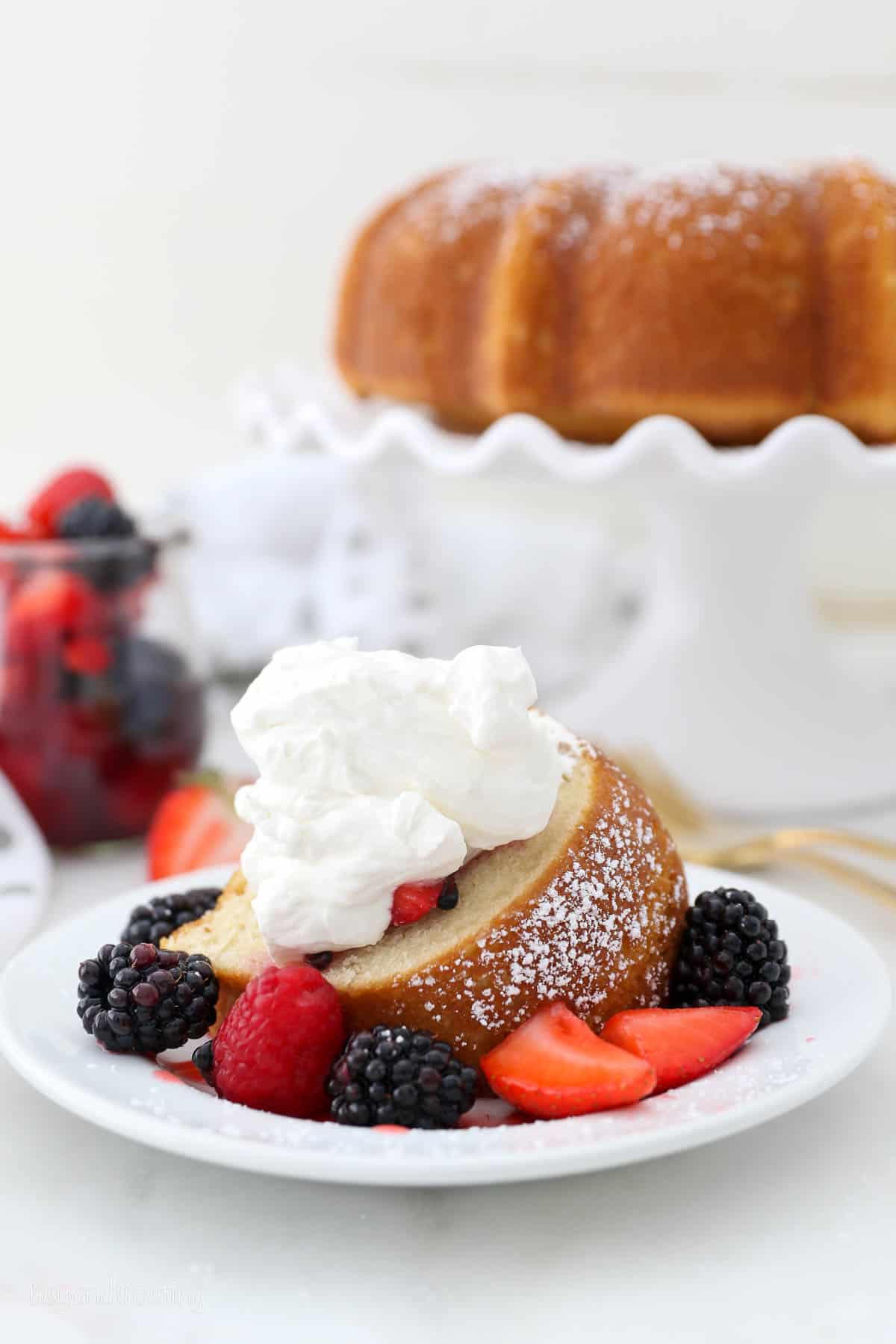 A slice of vanilla bundt cake on a white plate, surrounded by macerated mixed berries and topped with whipped cream, with the rest of the cake on a cake stand in the background.