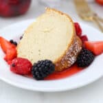 A slice of vanilla bundt cake on a white plate, surrounded by macerated mixed berries and topped with whipped cream.