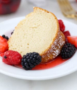 A slice of vanilla bundt cake on a white plate, surrounded by macerated mixed berries and topped with whipped cream.