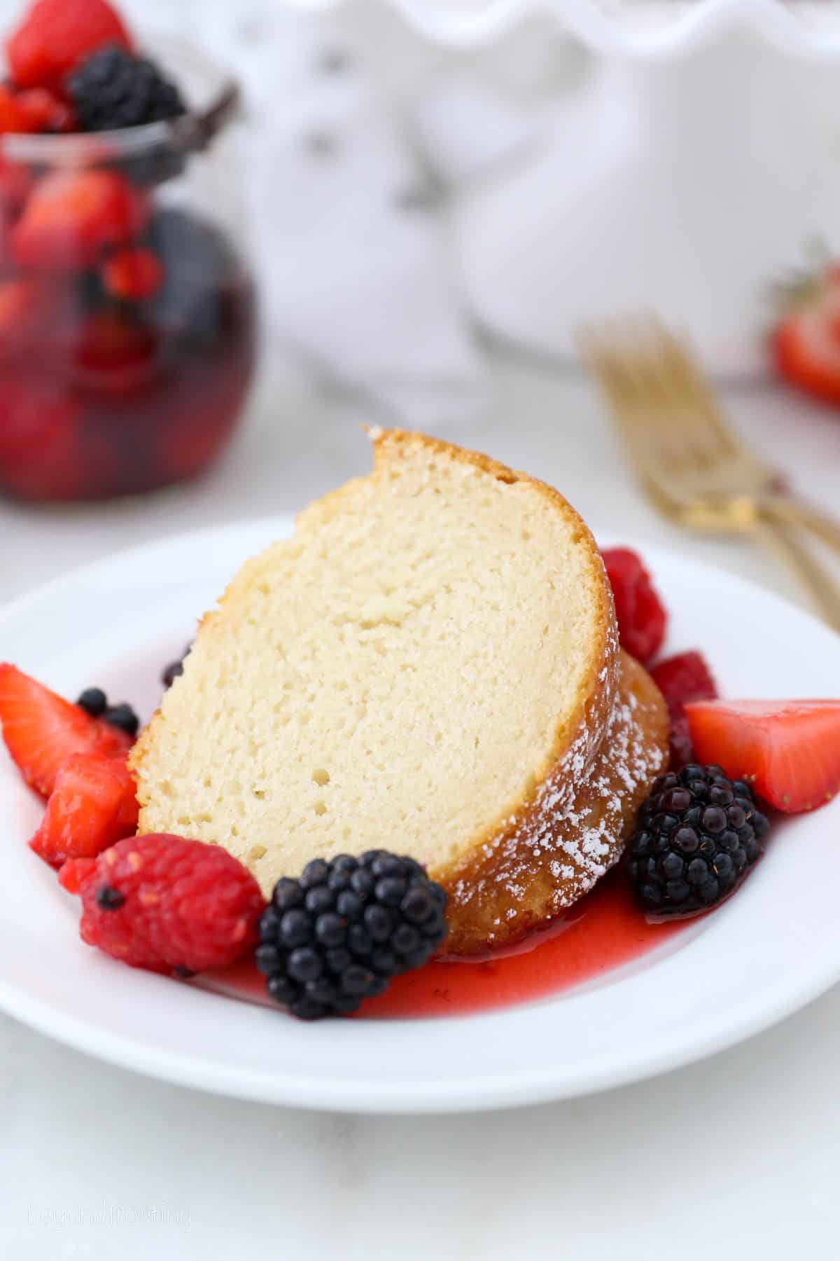 A slice of vanilla bundt cake on a white plate, surrounded by macerated mixed berries and topped with whipped cream.