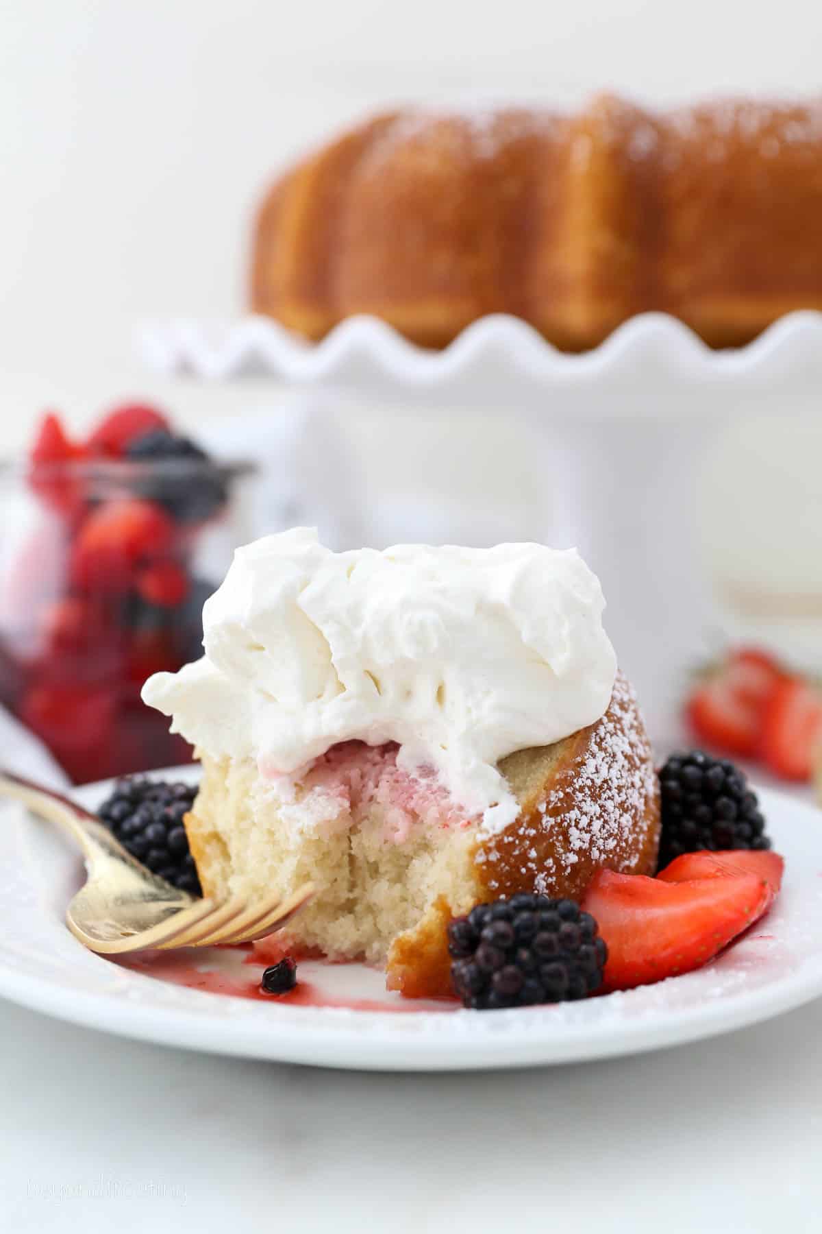 A slice of vanilla bundt cake on a white plate with a forkful missing, surrounded by macerated mixed berries and topped with whipped cream, with the rest of the cake on a cake stand in the background.