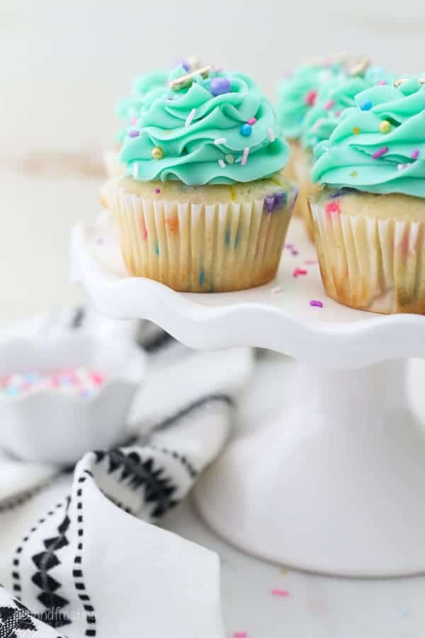 A small white cake stand with gorgeous funfetti cupcakes is draped with a white and black decorative napkin