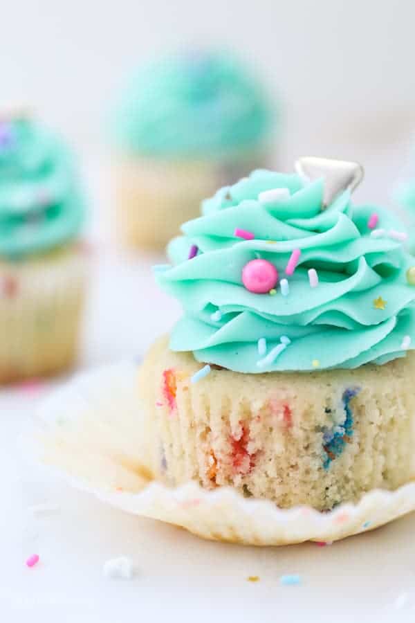 A closeup shot of a gorgeous funfetti cupcake with the wrapper pulled down and frosted with a light teal color frosting and garnished with colorful sprinkles