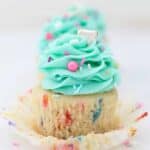 A gorgeous funfetti cupcake with the wrapper pulled down and frosted with a light teal color frosting and garnished with colorful sprinkles