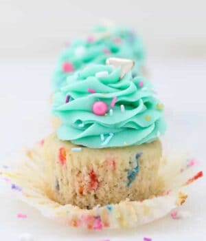 A gorgeous funfetti cupcake with the wrapper pulled down and frosted with a light teal color frosting and garnished with colorful sprinkles
