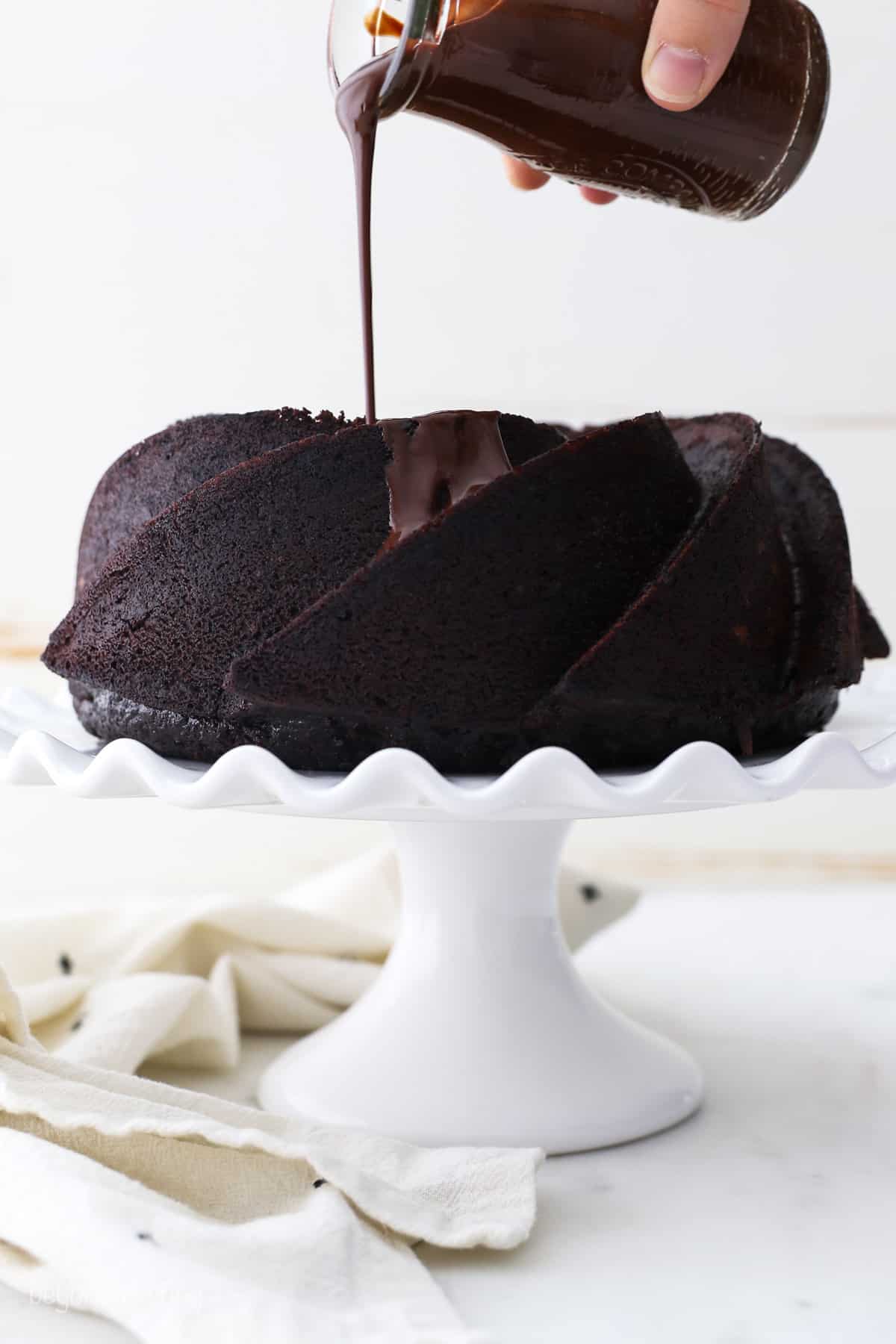 A hand pouring chocolate ganache over a chocolate bundt cake on a white cake stand.