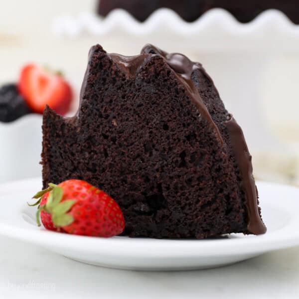 A slice of chocolate bundt cake next to a strawberry on a white plate.