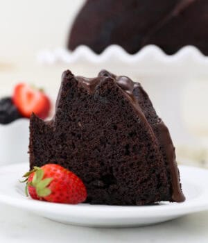 A slice of chocolate bundt cake next to a strawberry on a white plate, with the rest of the cake on a cake stand in the background.