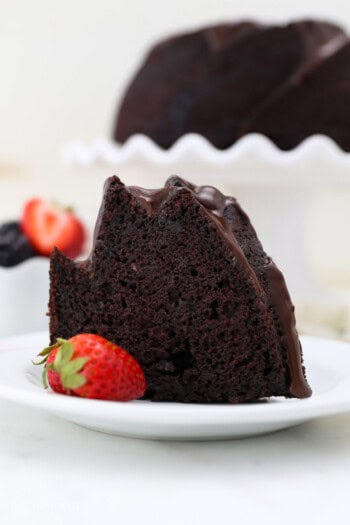 A slice of chocolate bundt cake next to a strawberry on a white plate, with the rest of the cake on a cake stand in the background.
