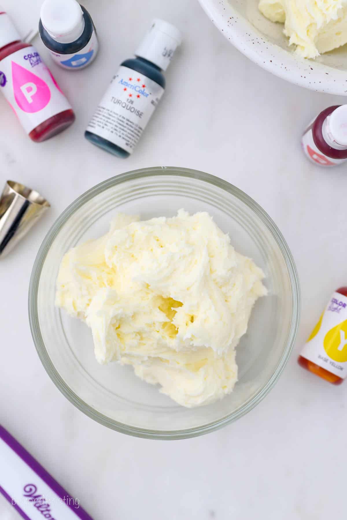 Overhead view of vanilla buttercream frosting in a glass bowl, surrounded by various bottles of gel food dye.