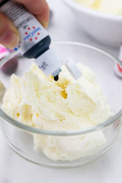 A hand squeezes a bottle of gel food dye over a bowl of vanilla buttercream.