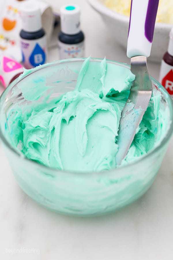 A glass jar of teal frosting with an angled spatula hanging out of the bowl and some color gels in the background