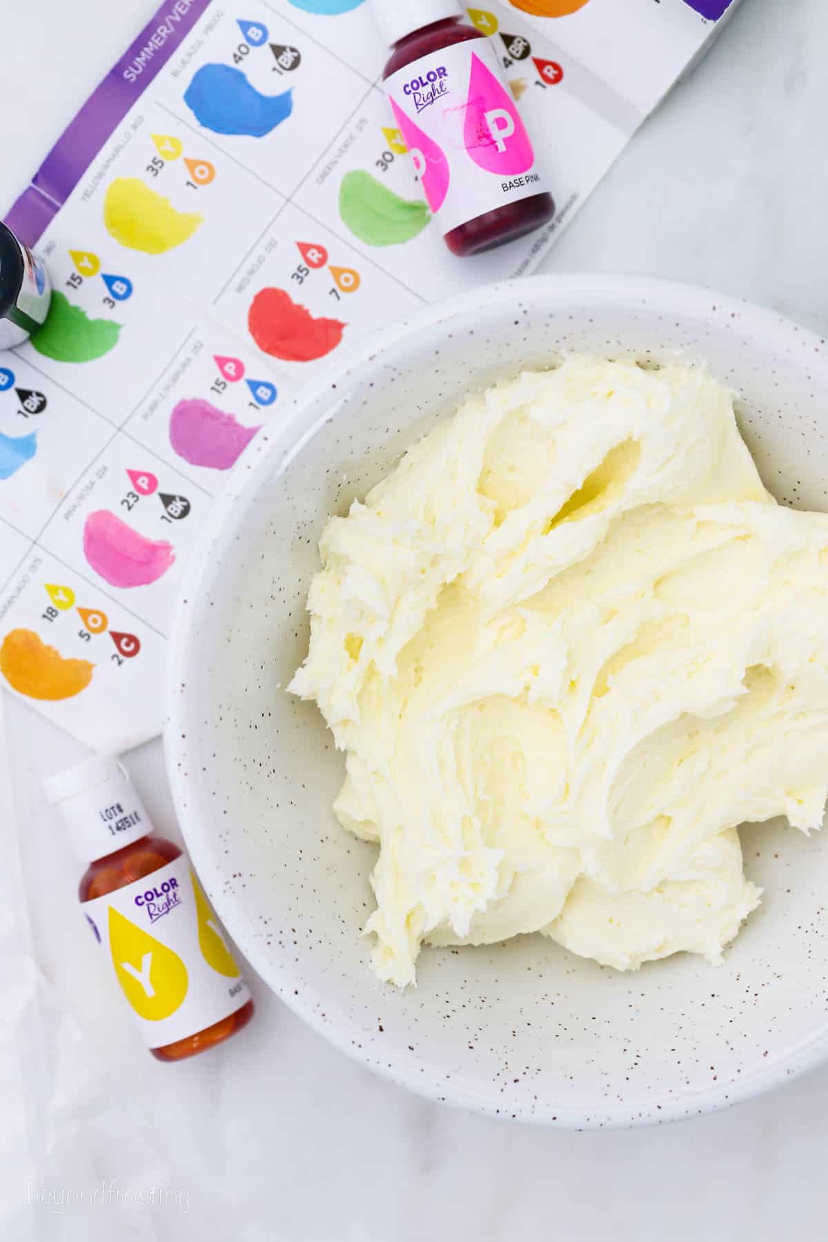 Overhead view of vanilla buttercream frosting in a bowl, next to two bottles of gel food dye and a color chart.