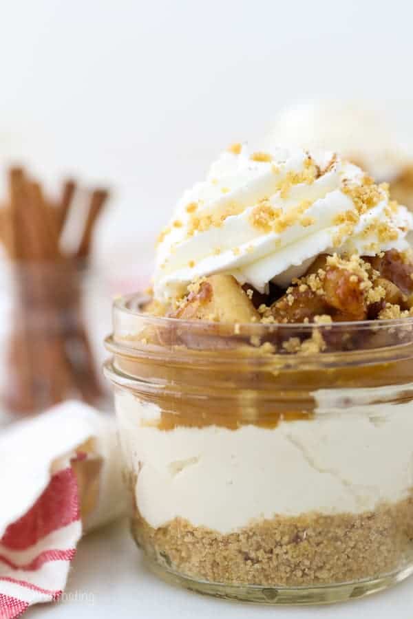 A close-up shot of a small glass jar filled with no-bake cheesecake filling, crust, apple pie filling, caramel sauce and whipped cream