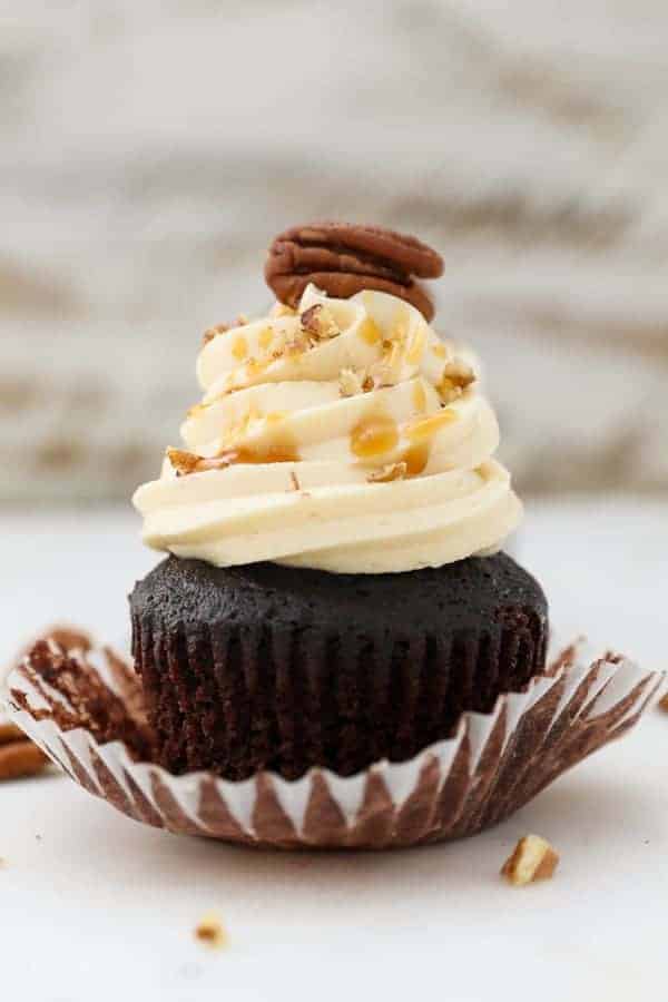 A chocolate cupcake with the wrapper pulled down, frosted with salted caramel buttercream and topped with a pecan