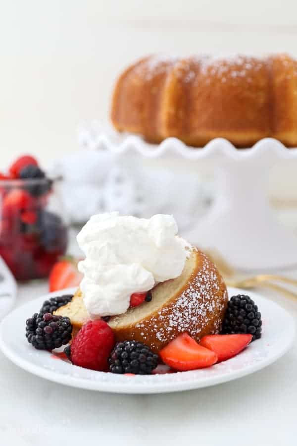 A pretty slice of vanilla cake bundt cake covered in berries and whipped cream. You can see the whole cake blurred out in the background