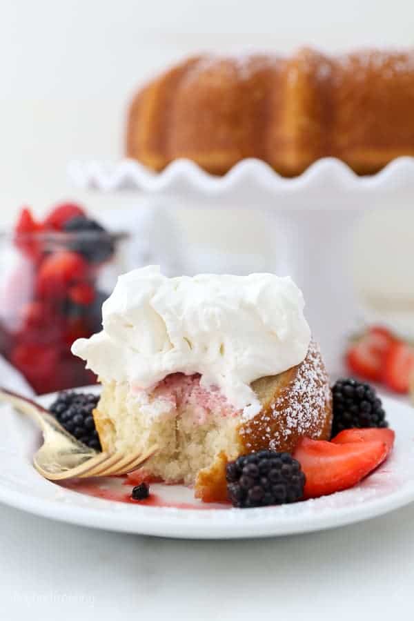 A slice of vanilla cake drizzled with berries and juice and topped with whipped cream, a gold fork is laying on the plate