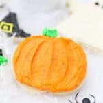 A pumpkin sugar cookie decorated with orange buttercream and sprinkles laying in a fake spider web