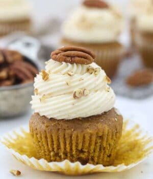 A pumpkin cupcake with the wrapper pulled down, it's frosted with a pretty swirled frosting and topped with a pecan.
