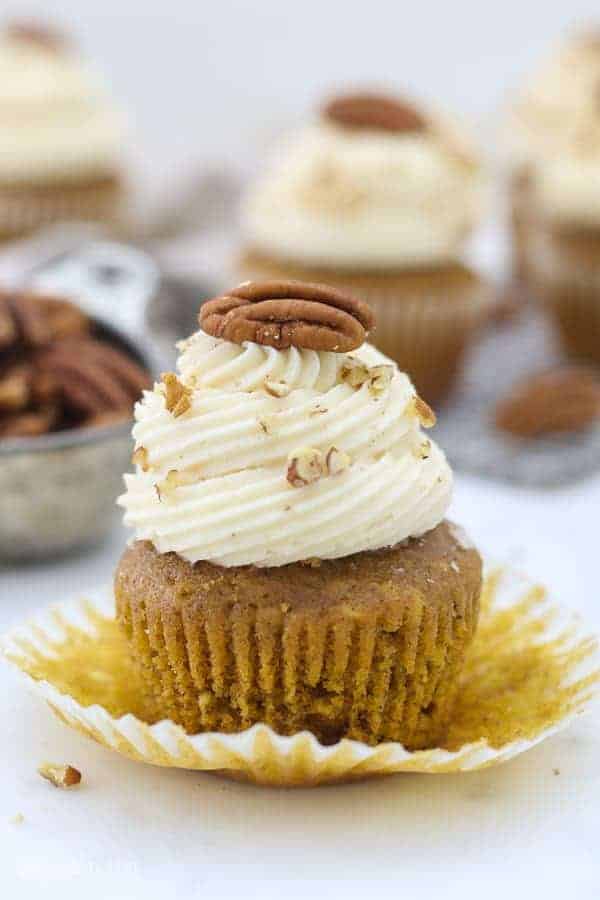 A pumpkin cupcake with the wrapper pulled down, it's frosted with a pretty swirled frosting and topped with a pecan.