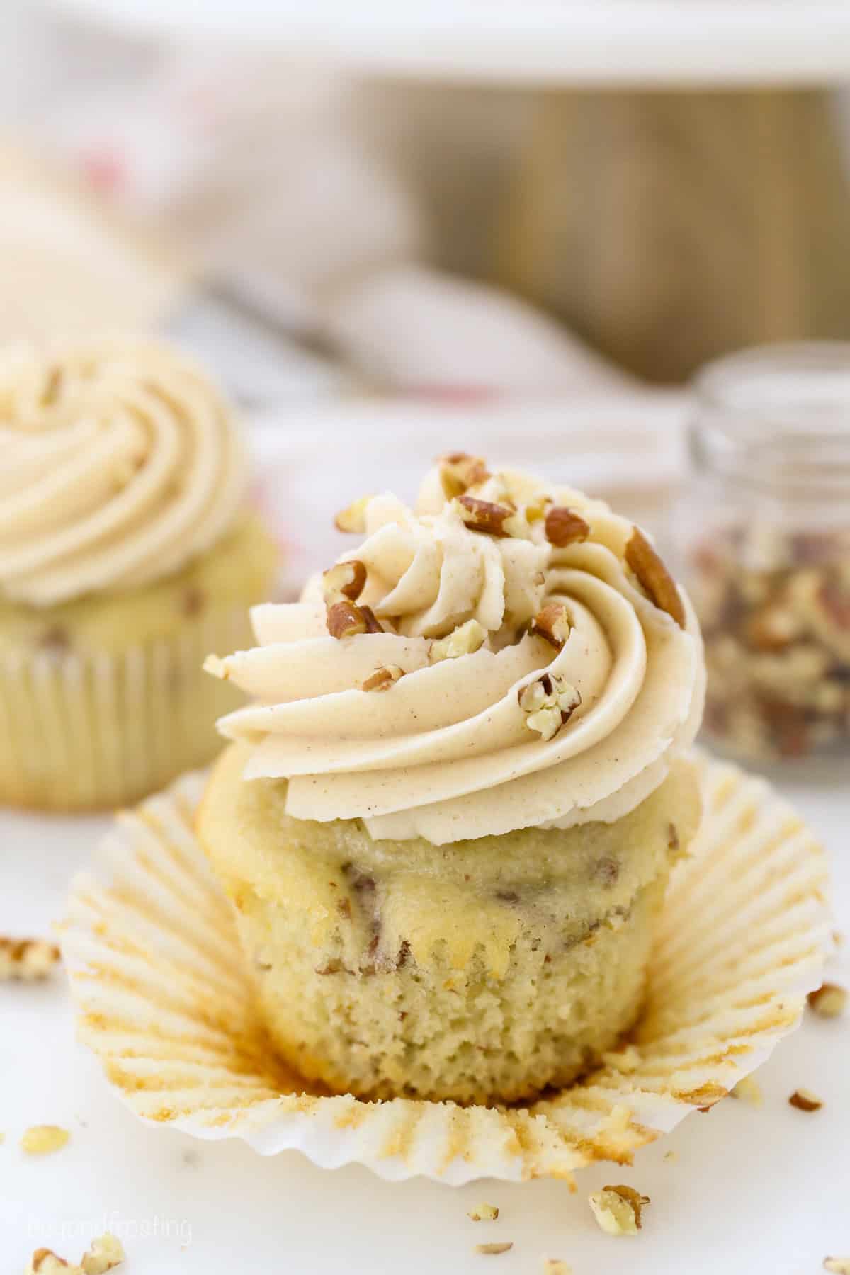 These homemade pecan cupcakes are filled with buttermilk, brown sugar, and buttery toasted pecans, finished with sweet maple buttercream. Close up of a pecan cupcake frosted with maple buttercream, partially unwrapped from a cupcake liner.