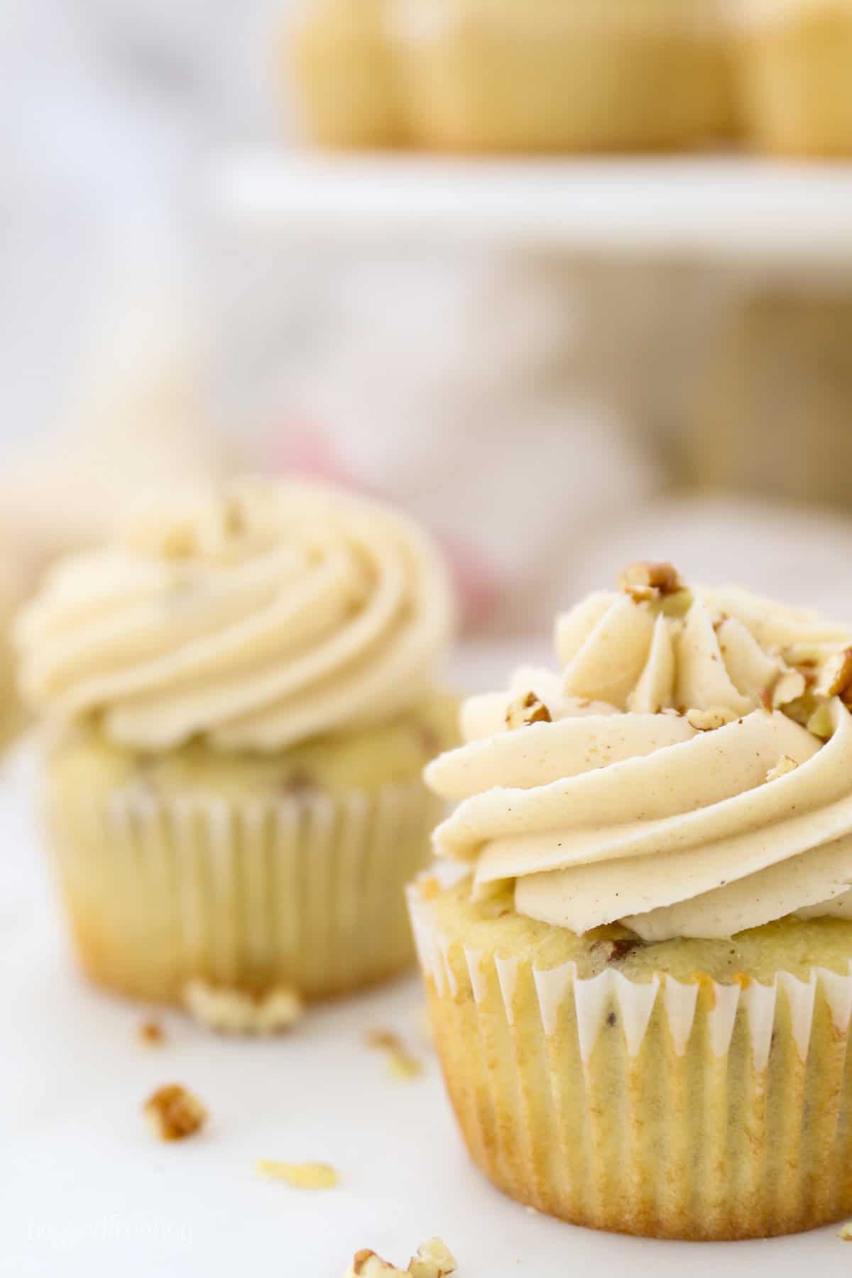 These homemade pecan cupcakes are filled with buttermilk, brown sugar, and buttery toasted pecans, finished with sweet maple buttercream. Close up of a pecan cupcake frosted with maple buttercream with a second cupcake in the background.