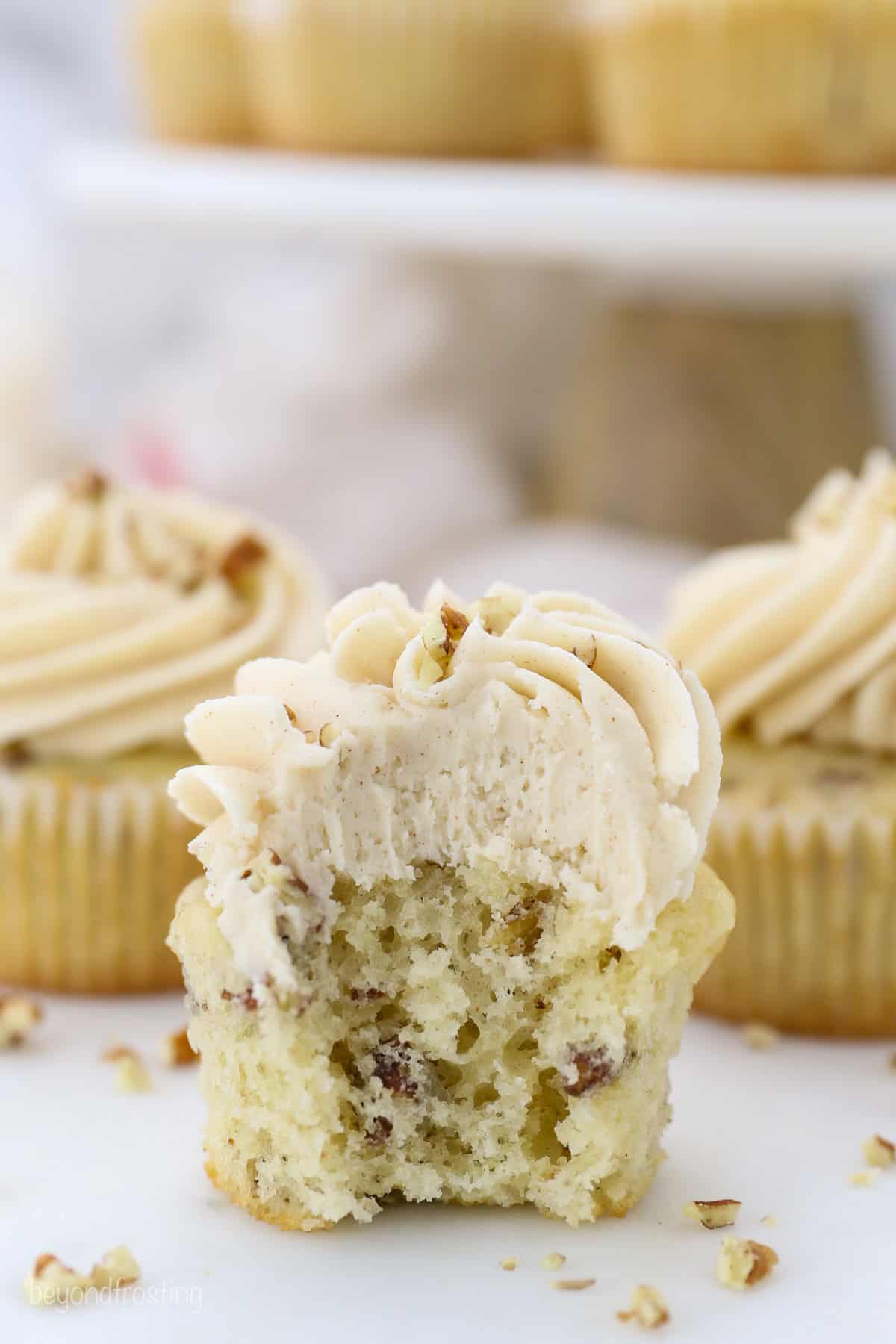 These homemade pecan cupcakes are filled with buttermilk, brown sugar, and buttery toasted pecans, finished with sweet maple buttercream. A frosted pecan cupcake with a bite missing, showing a tender, pecan-filled crumb.