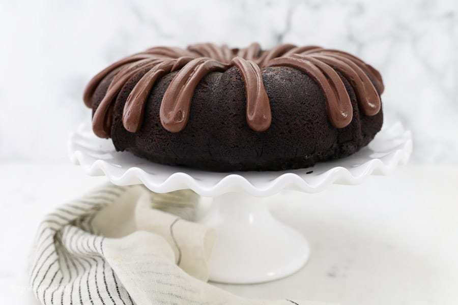 A chocolate bundt cake sitting on a white ruffled cake stand with a chocolate cream cheese frosting