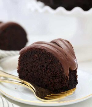 A slice of chocolate cake with a chocolate frosting is sitting on a white plate with 2 gold forks on it