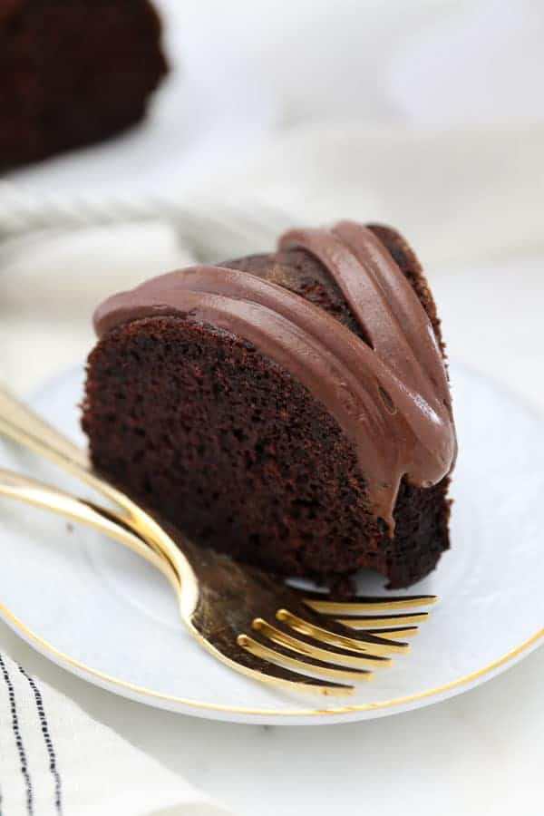 A overhead shot of slice of chocolate cake with a chocolate frosting is sitting on a white plate with 2 gold forks on it