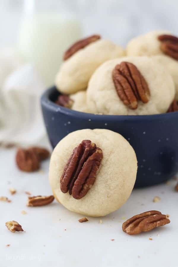 A sugar cookie with a pecan on top leaning up against a small blue bowl filled with cookies