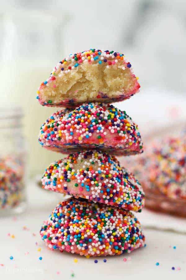 4 cream cheese cookies, rolled in rainbow sprinkles and stacked on top of each other, the top one has a bite taken out of it to show you the center of the cookie.