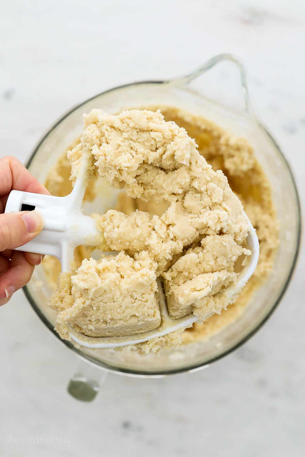A hand holding a stand mixer attachment filled with sugar cookie dough, with a bowl of cookie dough in the background.