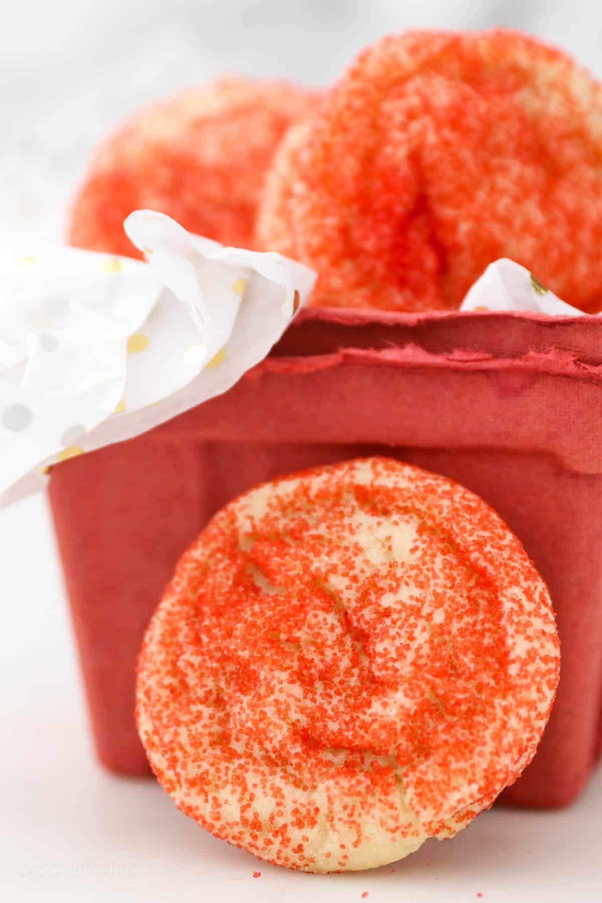 A soft sugar cookie coated in red sanding sugar leaning against a red carton of decorated sugar cookies.