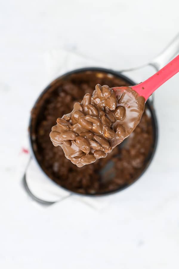 A red spatula is sitting above a saucepan with a scoop of melted chocolate and peanuts on it